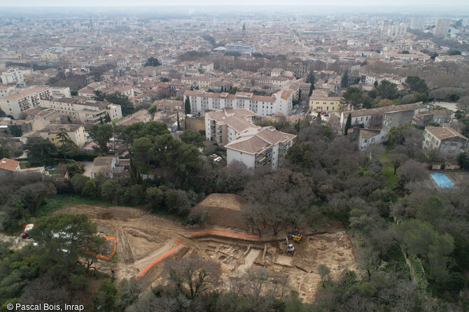 Vue aérienne du site avec les Arènes en arrière-plan. De riches vestiges d’un quartier urbain mis en place dès la Protohistoire (au IVᵉ siècle avant notre ère) et évoluant jusqu’à la fin du IIe siècle de notre ère ont été mis au jour de part et d'autres de la voie, à Nîmes (Gard), en 2021.